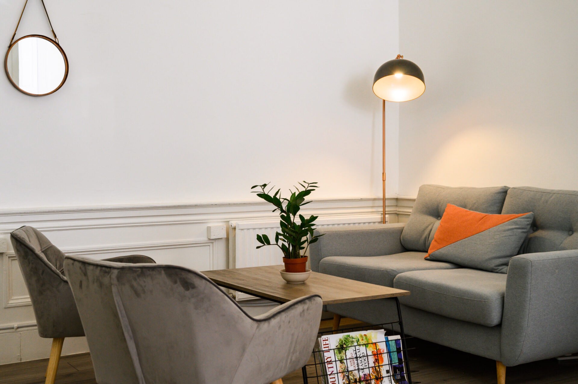 A cozy living room with a gray sofa and two velvet chairs around a wooden coffee table. A potted plant sits on the table, with a magazine rack below. A floor lamp and round mirror adorn the white walls. An orange pillow adds a pop of color.