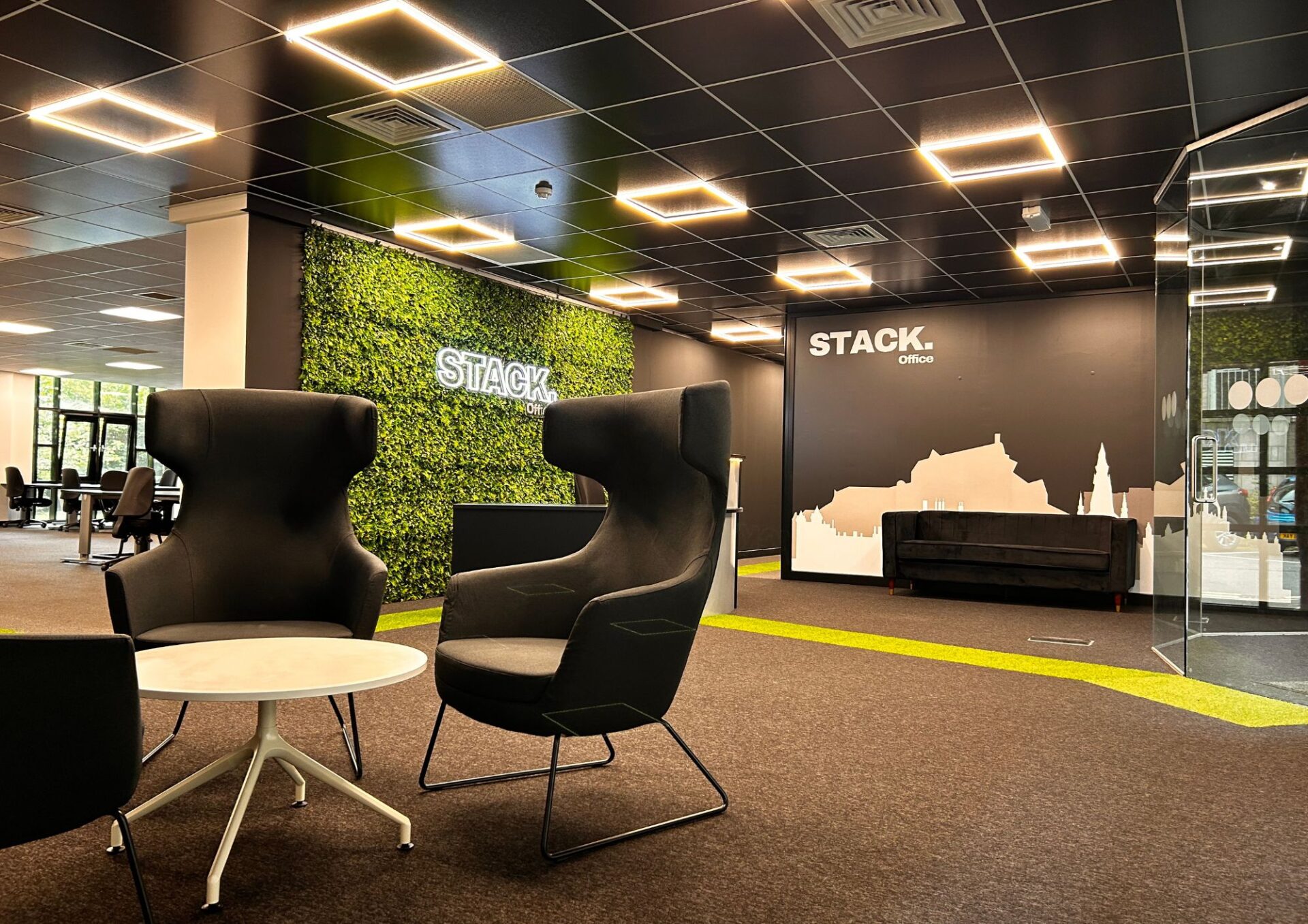 Discover STACK. Office 1 Papermill Wynd, where modern design meets functionality. This space boasts sleek black chairs around a round table, a captivating greenery wall, and a cityscape silhouette beneath geometric lights on the black ceiling.