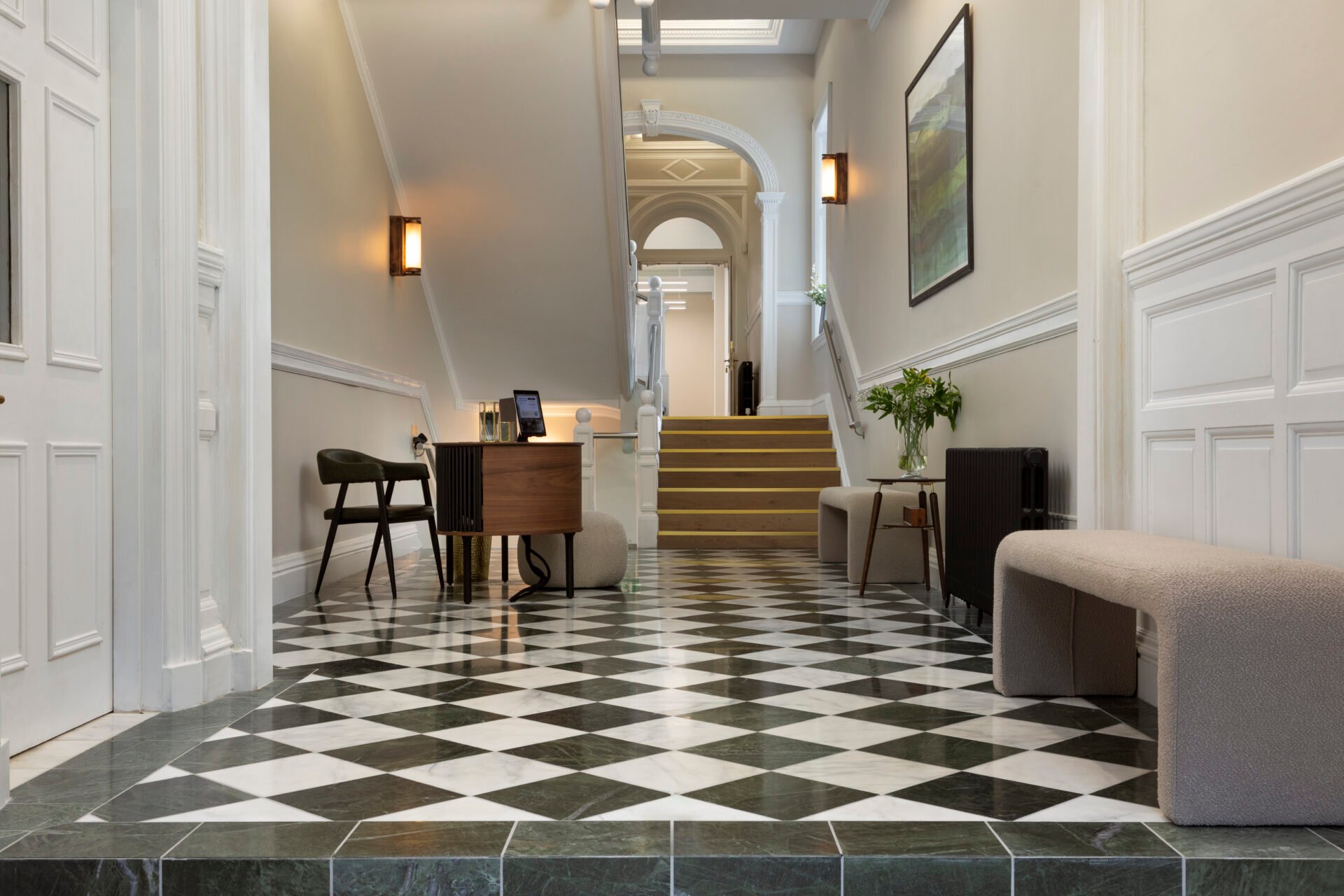 Located at 22 Queen Street, a hallway with a black and white checkered floor leads to a staircase. The walls boast white wainscoting, while a wooden desk and chairs sit beneath framed artwork. Soft lighting gently illuminates the inviting space.
