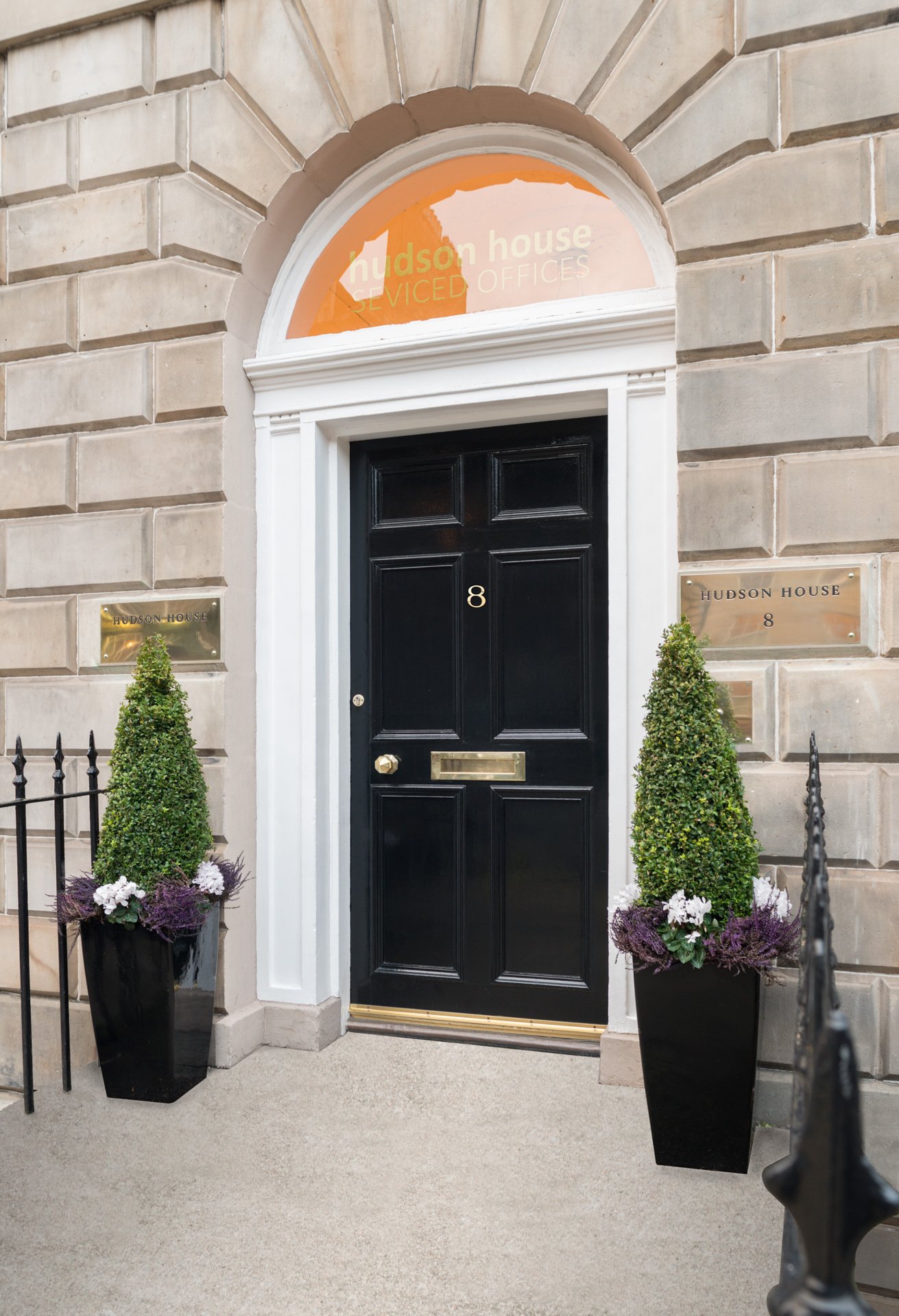 A black door marked with the number 8 is set within a stone facade, leading to Hudson House. Two tall potted plants flank the doorway, and a brass plaque adorns the wall. An arch above the door features the words serviced offices.