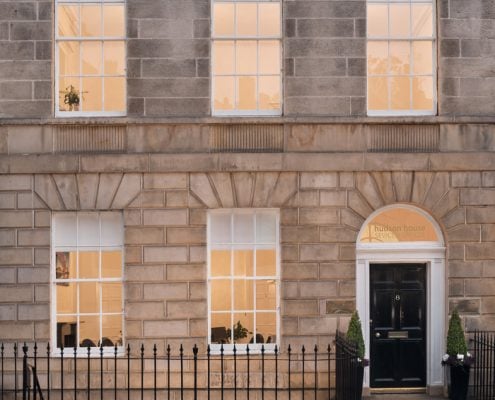 The elegant exterior of Hudson House features a classic stone facade with large windows and a black door flanked by charming potted plants. Above the entrance, a small arch window showcases the name, while a black iron fence gracefully borders the front.