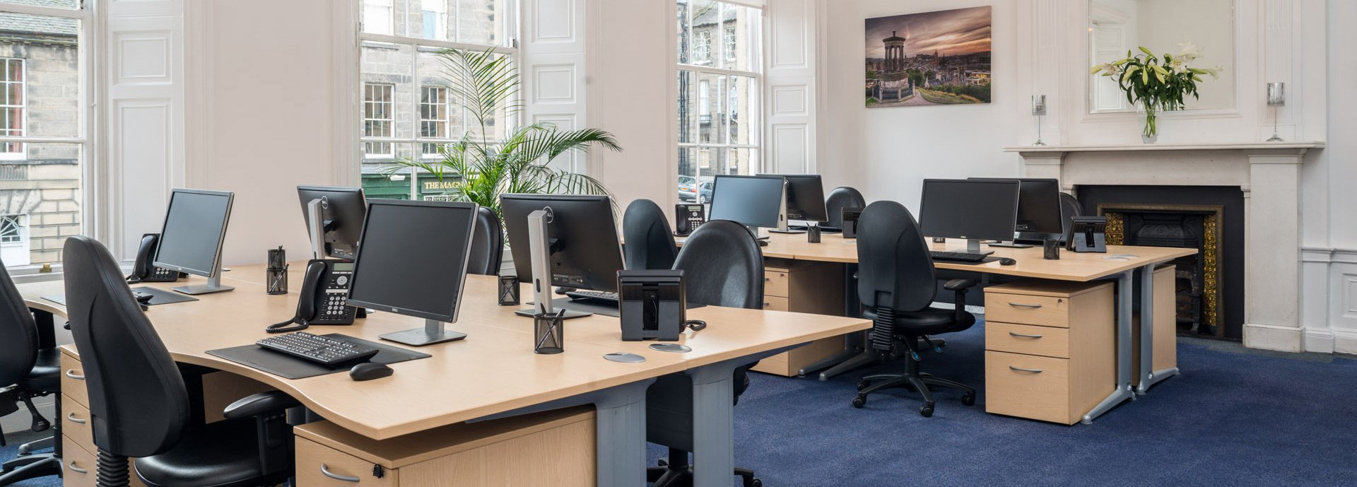 A modern office in Hudson House features multiple workstations arranged in an open layout. Each desk boasts a computer monitor and office chair. The room is accented by large windows, a plant, wall painting, and a fireplace adorned with flowers. Navy carpet and wood furniture complete the ambiance.