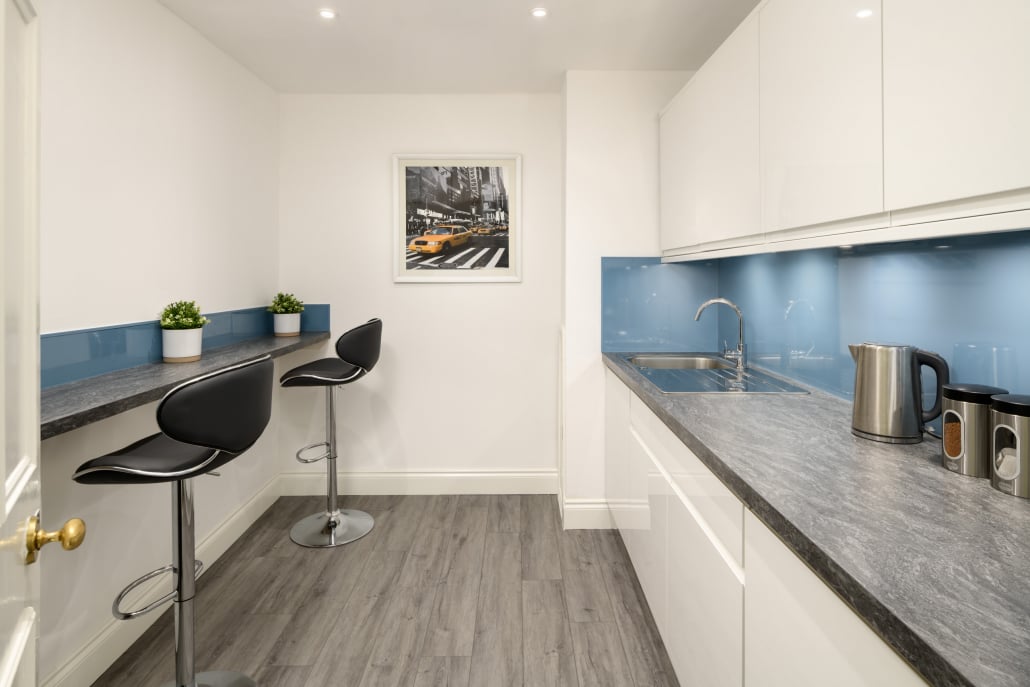 Modern kitchen at Hudson House featuring white cabinets, blue backsplash, and dark countertops. A sink, kettle, and mugs are on the right. Two black bar stools sit by a narrow counter on the left under a picture of a city street. Light wood flooring throughout enhances the space.