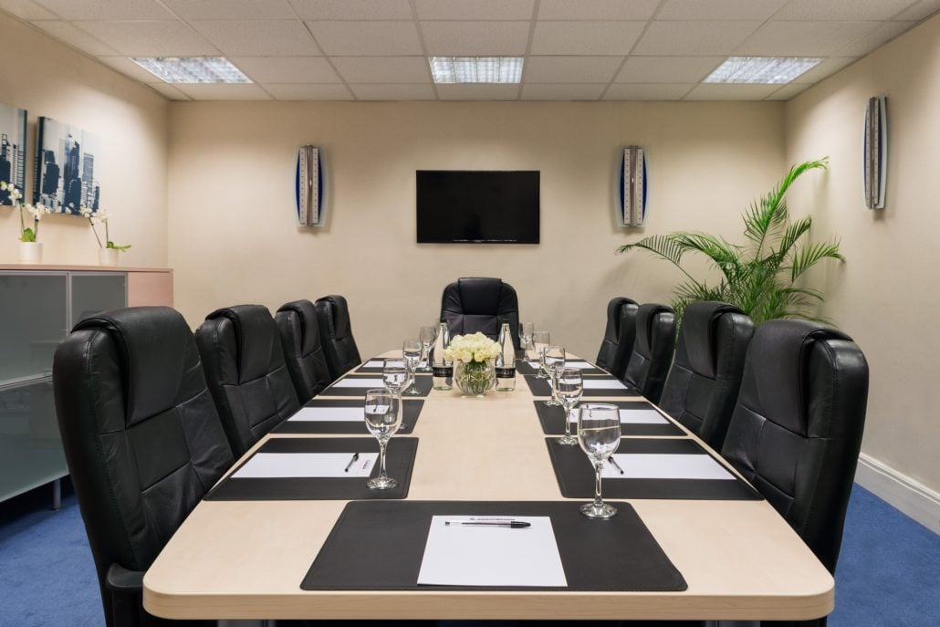 The conference room at Hudson House features a long wooden table surrounded by black leather chairs. Each setting includes a notepad, pen, water glass, and coaster. A TV is mounted on the wall, and a potted plant graces the corner under soft ceiling lighting.