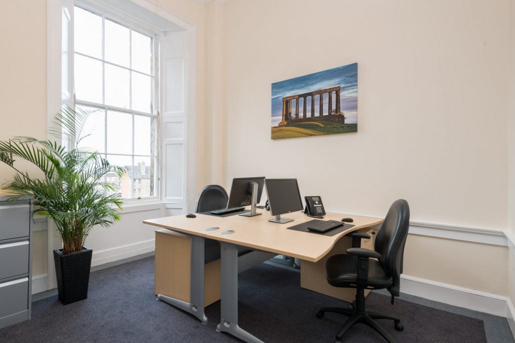 A tidy office in Hudson House features a large window, two office chairs, a wooden desk equipped with computers and a phone, a potted plant in the corner, and a picture of a classical building with columns on the wall.