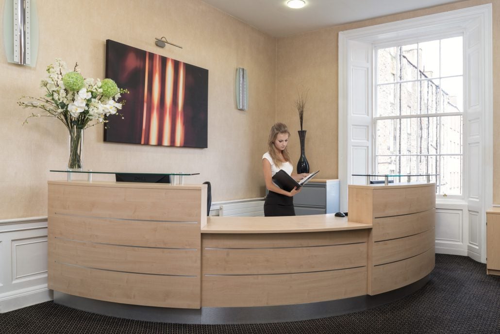 A woman stands behind a curved wooden reception desk at Hudson House, immersed in a book. The room features beige walls, a large window, and modern art on the wall. A vase with white flowers and green foliage adorns the counter, enhancing the serene ambiance.