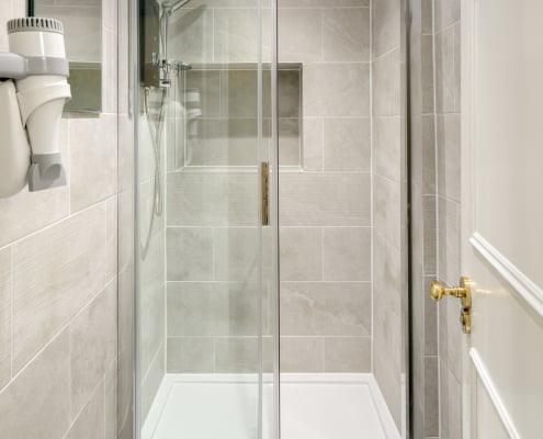 A modern bathroom in Hudson House showcases a glass-enclosed shower with light beige tiles. A built-in shelf hosts a sleek showerhead, while a wall-mounted hairdryer adorns the left side. The white door, accented by its gold handle, is slightly ajar.