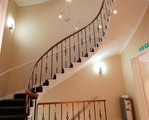 In Hudson House, a spiral staircase with a dark wooden handrail and metal balusters ascends in a well-lit hallway. Warm beige walls are adorned with modern wall sconces, and theres a green exit sign above a door on the upper right.
