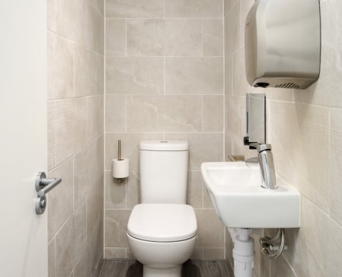 The small bathroom at Hudson House boasts beige tiled walls, a white toilet, and a wall-mounted sink with a metallic faucet. A hand dryer rests above the sink, while the toilet paper holder is on the left wall, complementing the partially open doors inviting nature.