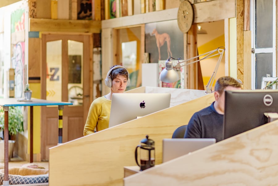 In the cozy Tribe Porty office with wooden elements, one person listens to music on headphones while working on an Apple computer, as the other uses a Dell. A coffee press graces the desk, and natural light fills the room with warmth and inspiration.