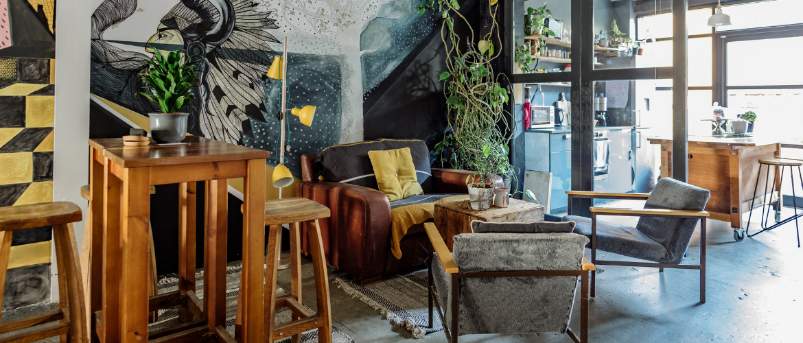 A cozy living room with a brown leather sofa and gray armchairs surrounding a wooden coffee table exudes the welcoming vibe of Tribe Porty. A bird mural adorns the wall, while indoor plants and a tall wooden stool highlight the modern kitchen in the background, all bathed in natural light.