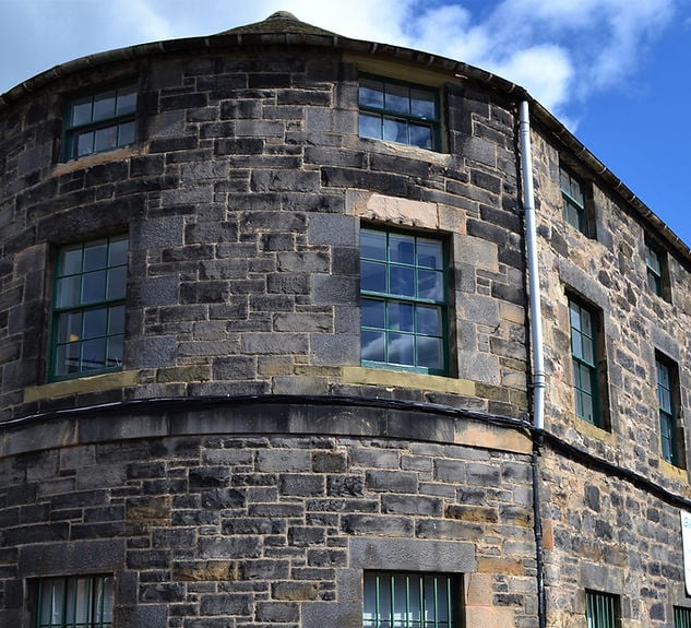 A rounded corner of an old stone building on Giles Street, with green-framed windows under a partly cloudy blue sky. The walls are dark bricks with lighter trim that echoes the artistic flair of the nearby studios.
