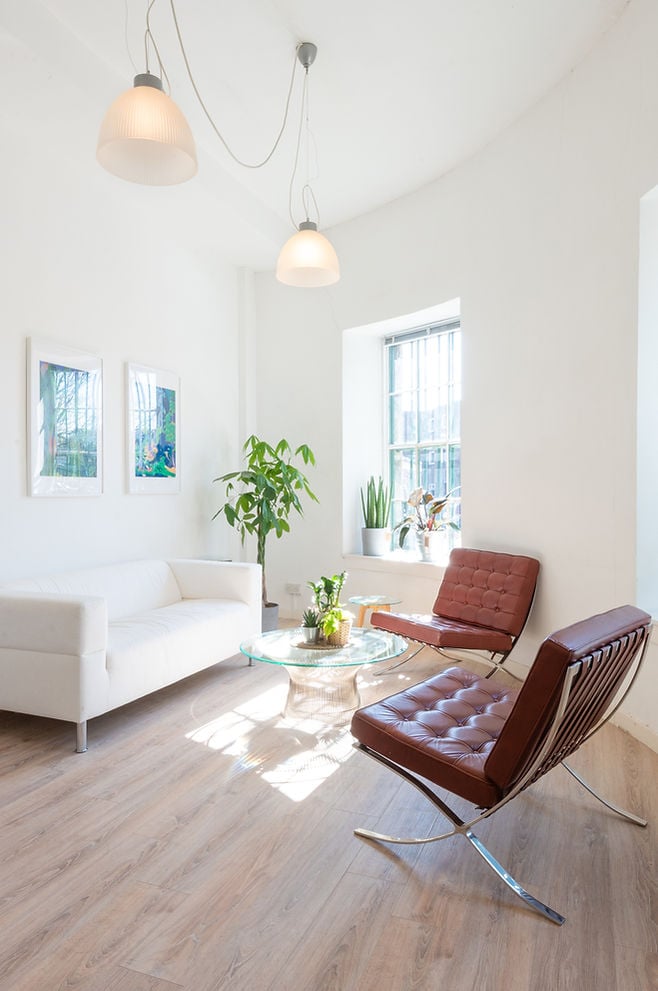 A bright, modern living room designed by Giles Street Studios features a white sofa, two brown leather chairs, and a glass coffee table. It boasts a light wooden floor, potted plants, and two framed artworks on the wall, all under elegant pendant lights.