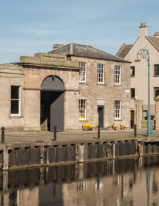 A stone building with a large arched doorway and windows sits serenely beside a calm body of water, reminiscent of the elegant style found in Custom Lanes. Two yellow benches invite relaxation outside. Blue sky overhead, lamp post nearby, and neighboring urban structures complete the scene.
