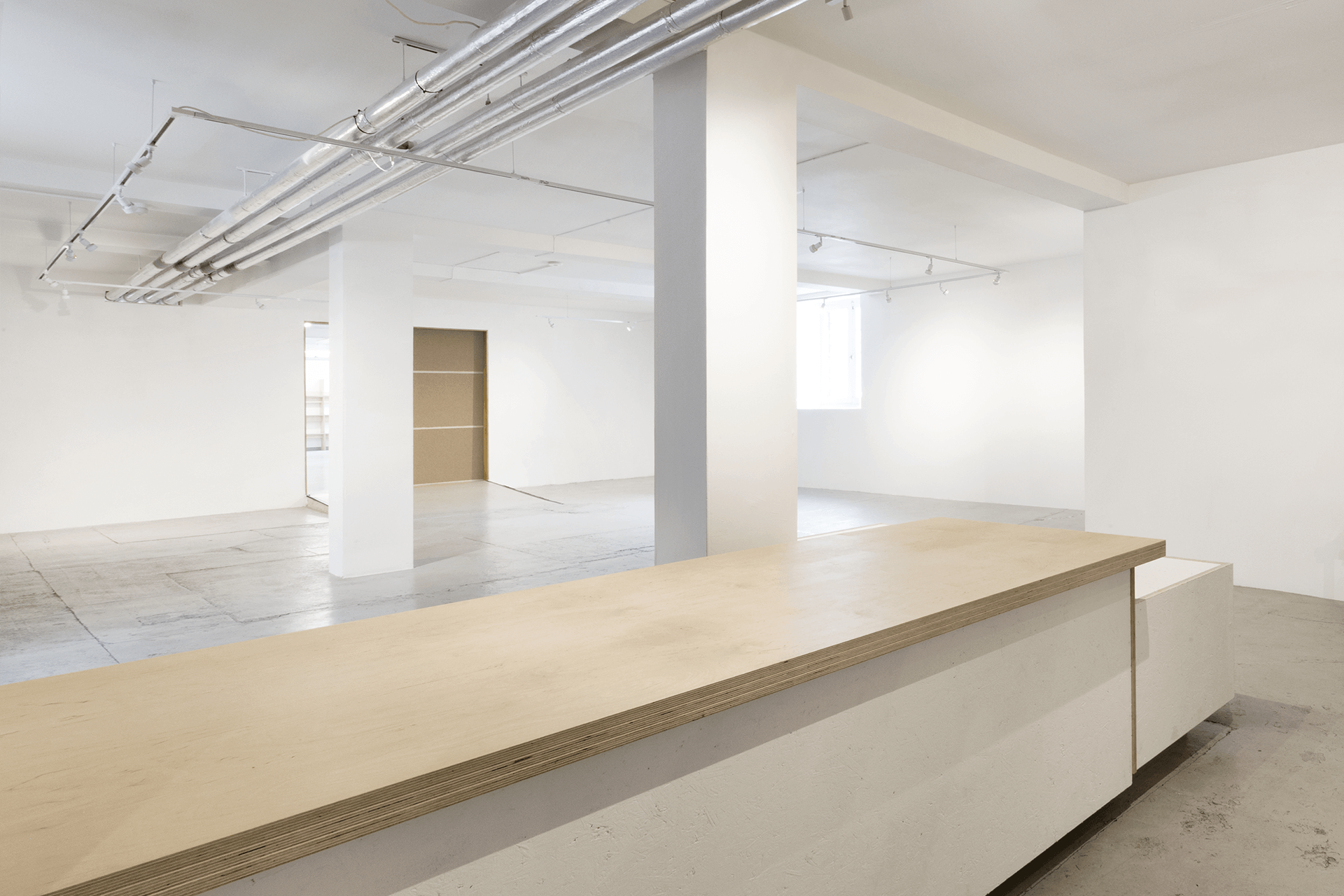 Spacious, empty gallery space with white walls and ceiling, exposed pipes, and concrete floors in Custom Lanes. A wooden counter is in the foreground. Natural light enters through a window in the back, highlighting the minimalist design.