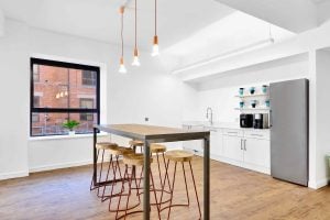 Modern kitchen with wooden floors and white walls. Features a tall dining table with four wooden stools, a set of hanging light bulbs, and a window offering a view of another building. A touch of foxglove decorates the white kitchenette with its fridge, coffee maker, and shelves.
