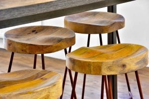 Four wooden stools with smooth, rounded seats and metal legs are arranged under a table, their presence echoing the charm of a rural kitchen. The grain and natural tones of the wood show through, like foxglove blossoms accenting an idyllic garden. The floor is made of light-colored wood.