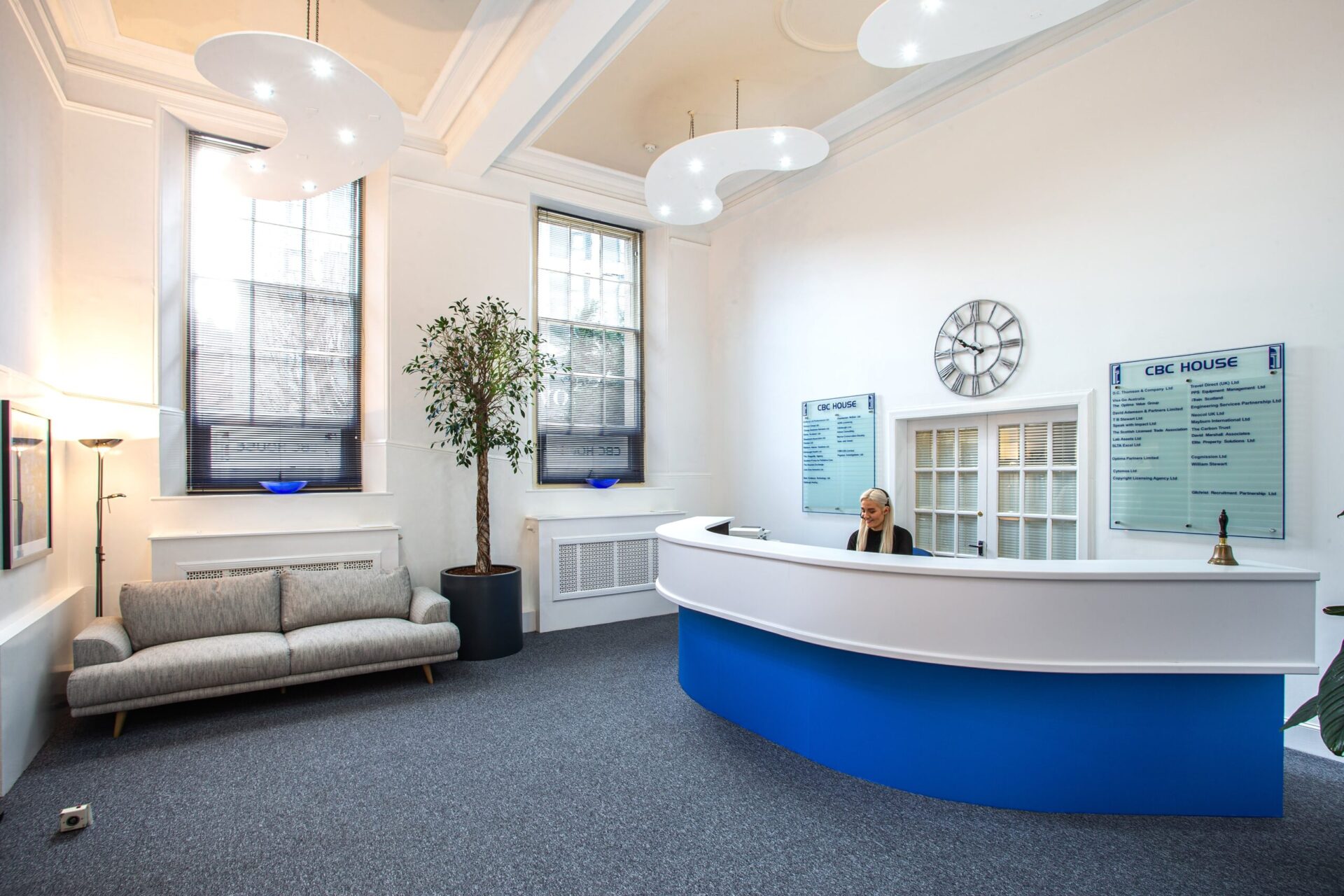 A modern, bright reception area at CBC House features a white and blue curved desk with a clock on the wall and a receptionist seated behind it. Large windows, a potted plant, and a gray couch elegantly complete the welcoming space.