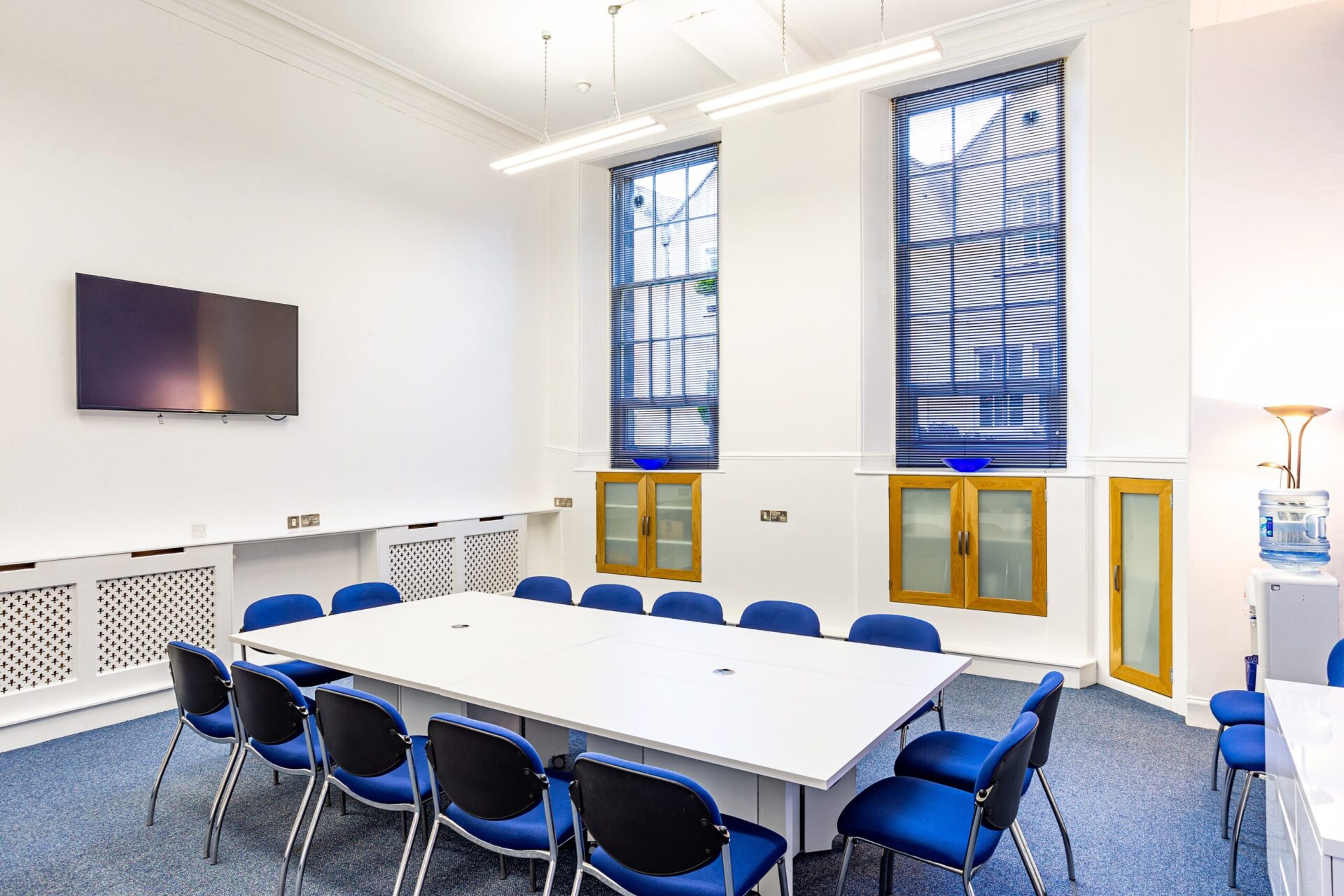 The modern CBC House conference room boasts a rectangular white table surrounded by twelve blue chairs. A wall-mounted TV graces one side, while tall windows with matching blue blinds line another. In the corner, a water cooler and lamp complement the brightly lit space.
