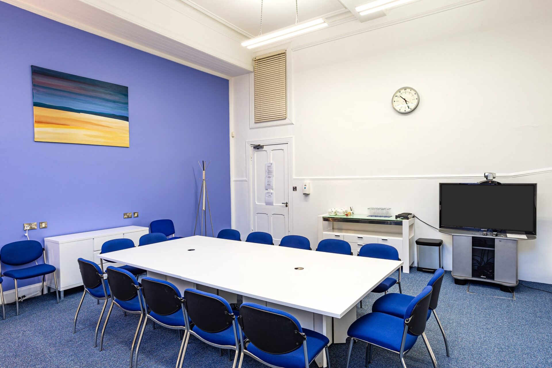The modern conference room at CBC House features a long white table surrounded by blue chairs. Blue walls host a colorful abstract painting, while a clock keeps time. A TV and snacks are neatly arranged on the white cabinet nearby, enhancing the inviting atmosphere.