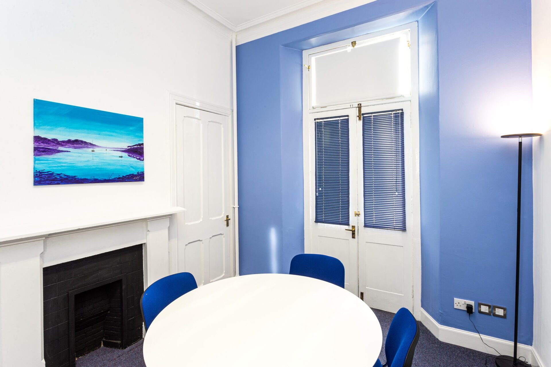 The CBC House boasts a bright room with blue walls, featuring a round white table surrounded by four blue chairs. A landscape painting graces the space above a closed fireplace, while two tall white doors, partially draped in blinds, stand beside a floor lamp.