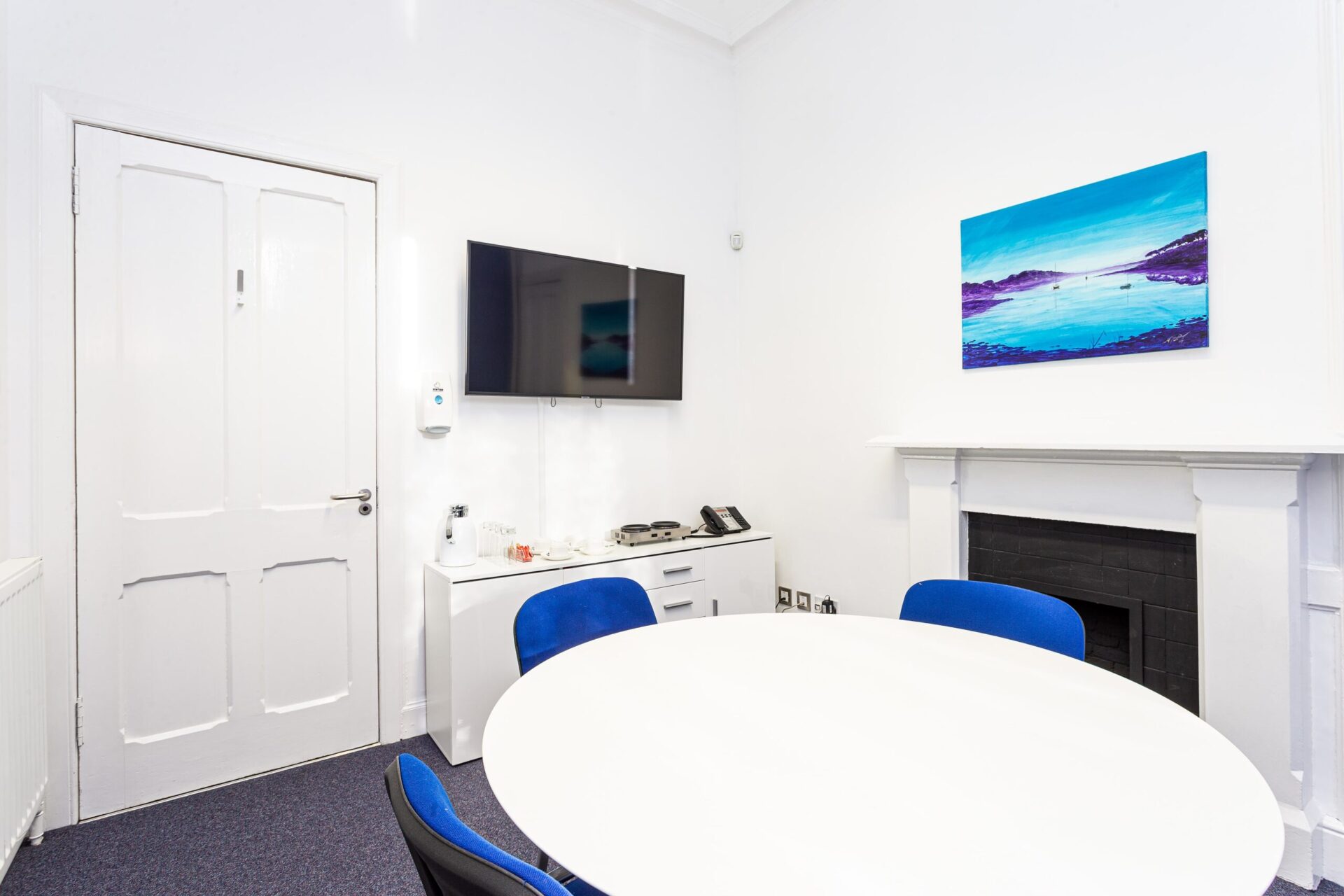 The CBC House conference room offers a cozy setting with a round white table and four blue chairs. A flat-screen TV, phone, and tea set grace the white cabinet. Above the mantelpiece, a vibrant landscape painting bursts in shades of blue and purple.