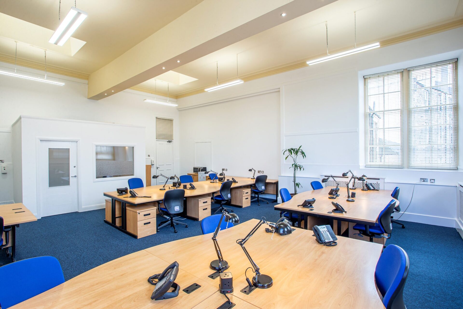 The spacious office at CBC House features blue carpet and wooden desks arranged in clusters. Surrounding each desk are blue chairs equipped with lamps and phones. Large windows on the right allow natural light, while white walls and ceiling lights enhance the bright ambiance.