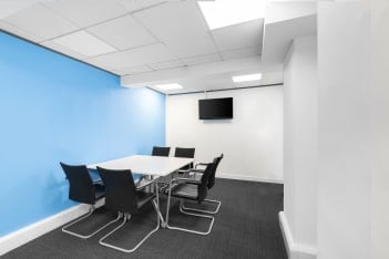 A modern conference room echoes the style of Princes Street, with a blue accent wall contrasting white surfaces. It features a rectangular white table and five black chairs. A flat-screen TV adorns the wall, while the dark gray carpet adds sophistication underfoot.