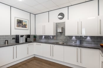 Modern kitchen with white cabinets, gray subway tile backsplash, and dark countertops. A wall clock is above, with a framed picture of Princes Street beside it. Stainless steel appliances and a small plant decorate the space. Brown wooden floor completes the look.