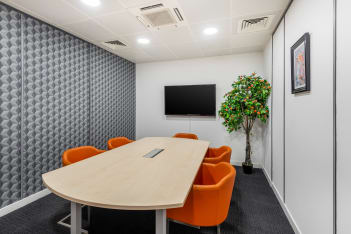 A modern conference room on Princes Street features a long wooden table, six bright orange chairs, and a large flat-screen TV mounted on the wall. A potted plant adds life, while textured gray panels and a framed picture enhance the aesthetic.