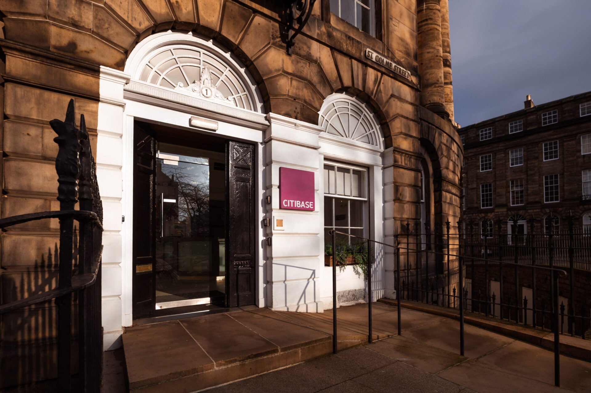 Entrance of a historic building with a label CITIBASE beside the door. The structure features large arched windows, stone columns, and a staircase. Sunlight casts shadows, emphasizing the architectural details. Another building is visible in the background.