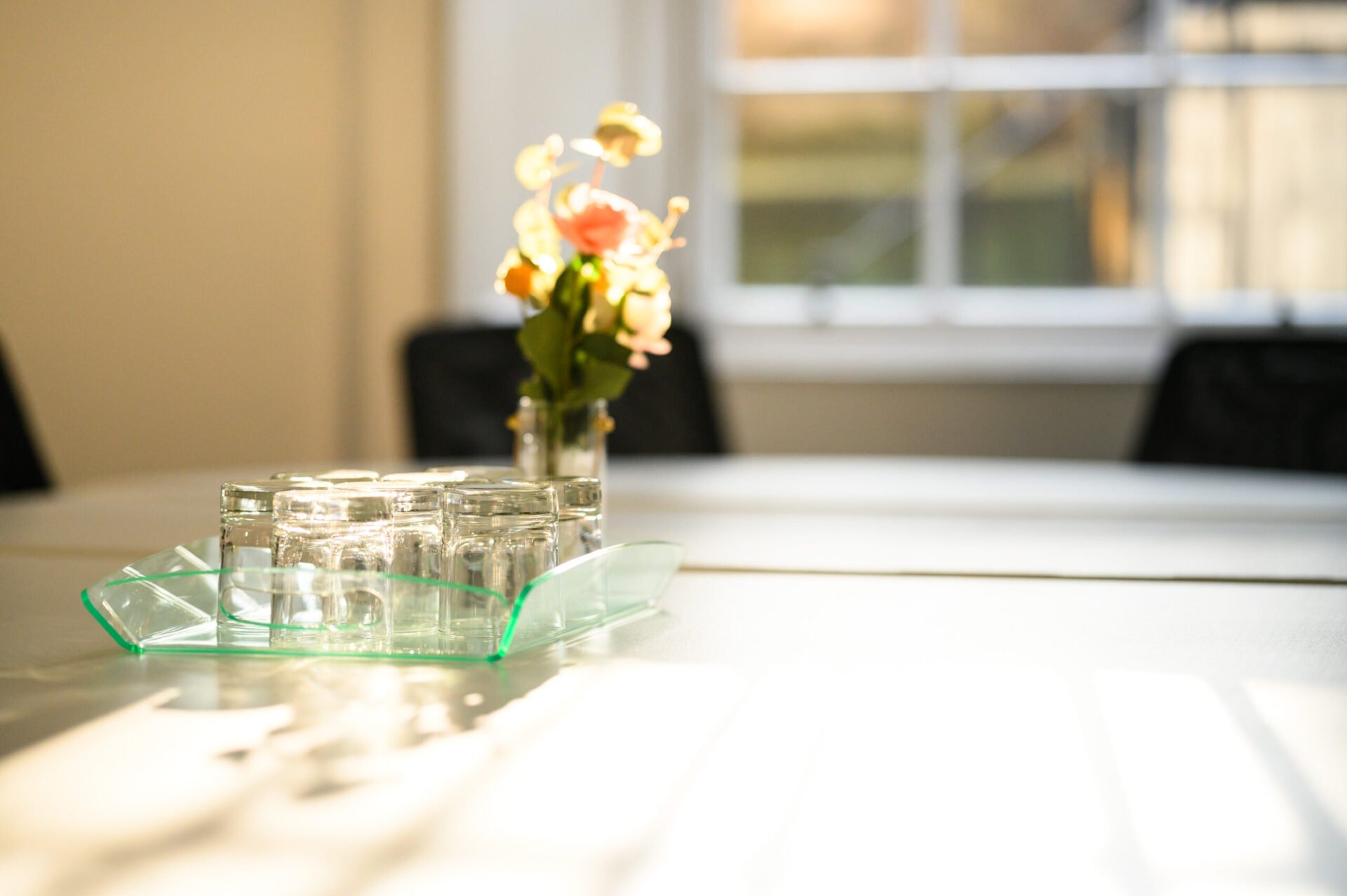 A sunlit conference table with a small glass vase holding flowers and a tray of neatly arranged empty glasses. Warm light streams in through a window, casting soft shadows on the table.
