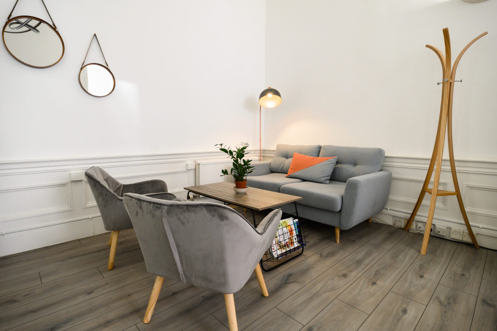 A modern living room with a gray sofa, two gray armchairs, and a wooden coffee table on a wooden floor. A floor lamp and plant are next to the sofa, with two circular mirrors on the white wall and a wooden coat rack nearby.