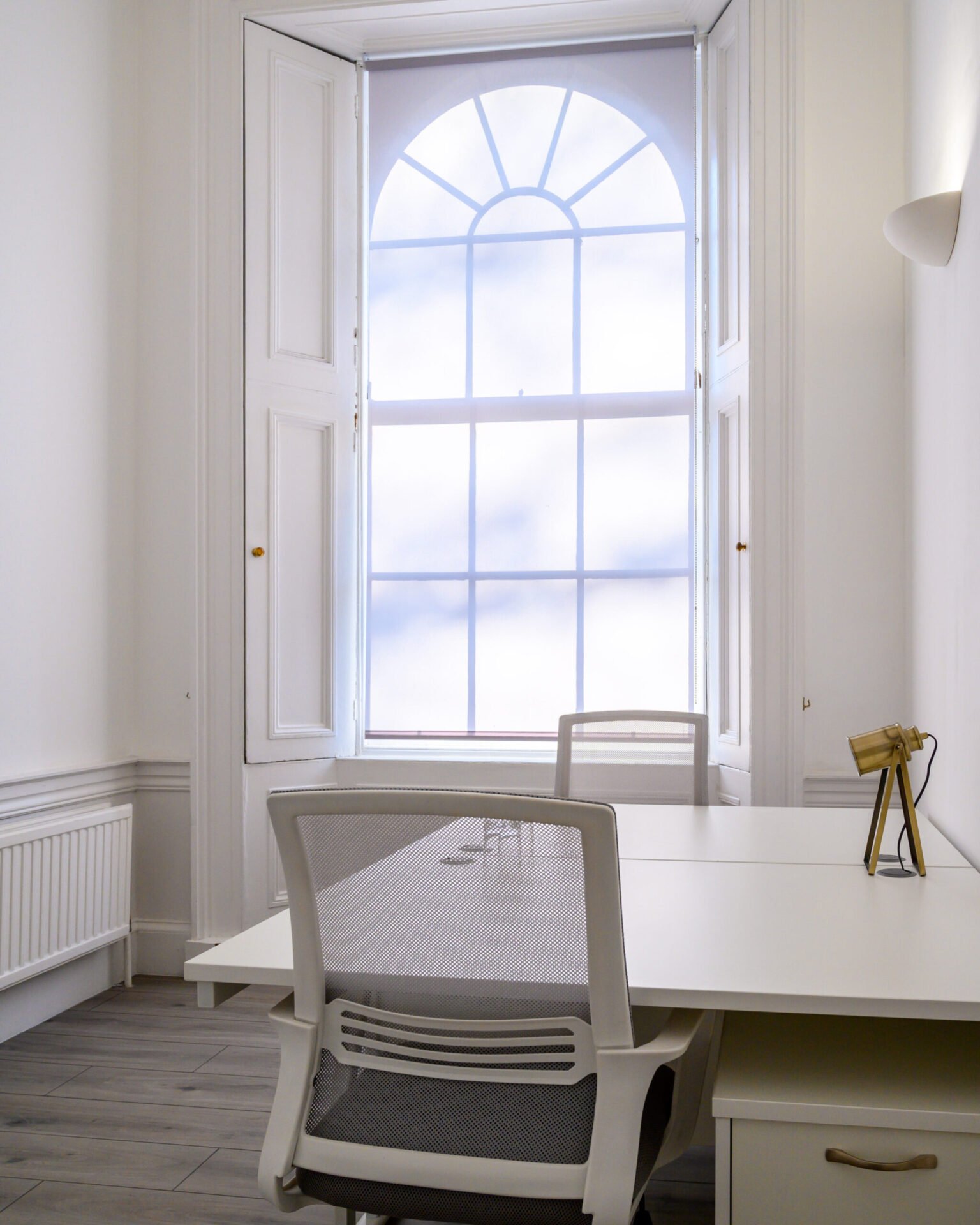 A bright office space featuring a white desk with a mesh office chair. The room has a large arched window with soft blue sky visible outside. The walls are white, and the floor is light gray wood. There is minimal decor, creating a clean aesthetic.