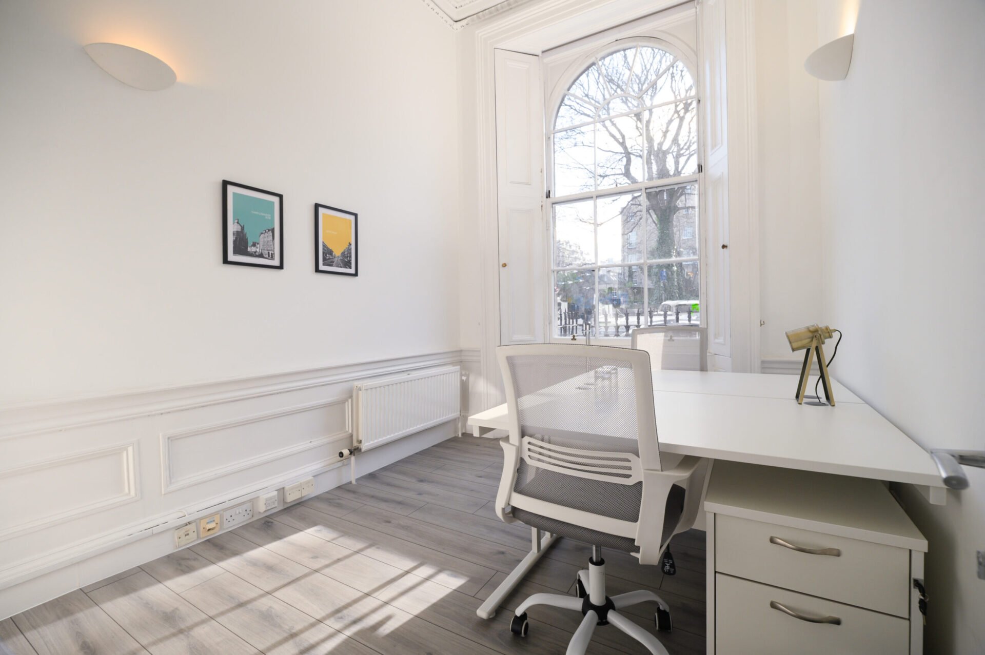 Minimalistic office with a white desk, ergonomic chair, and large window. Sunlight streams through, illuminating the light wood floor. Two framed prints hang on the white wall, and a small vase sits on the desk.