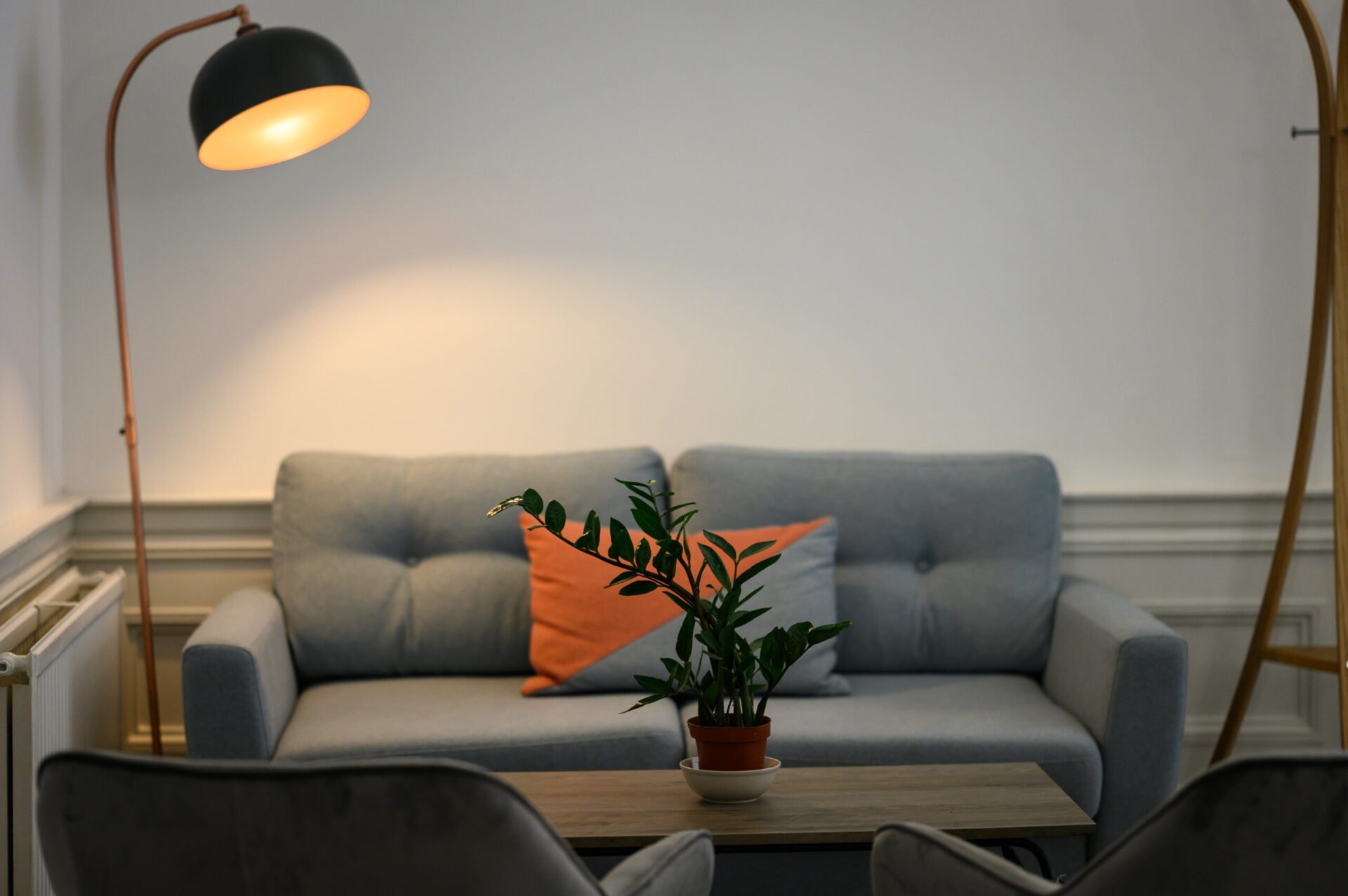 A cozy living room with a gray sofa featuring an orange throw pillow. A tall floor lamp casts a warm light over the area. A coffee table with a potted plant sits in front of the sofa, and two chairs are partially visible.