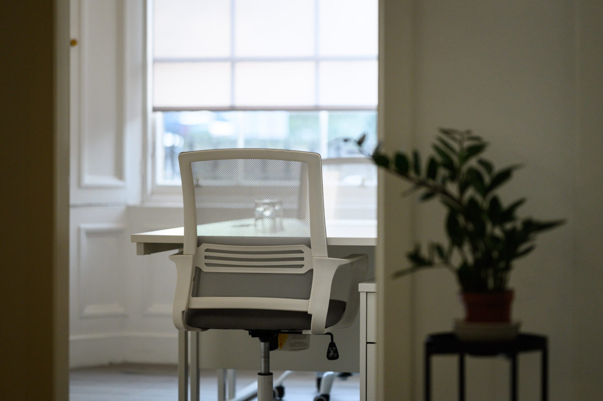 A modern office chair in front of a desk with soft natural light streaming through a large window. A potted plant sits on a small table to the right, adding a touch of greenery to the neutral-toned room.
