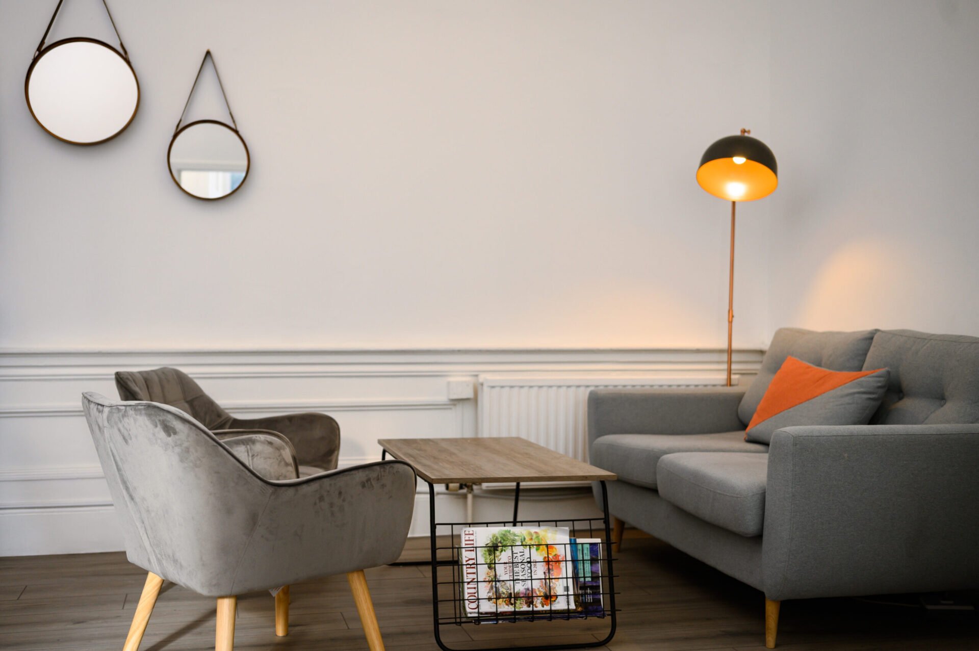 A modern living room with a gray sofa, a wooden coffee table with magazines, and two gray armchairs. Two round mirrors hang on the white wall. A floor lamp with a warm light stands nearby. An orange pillow accents the sofa.