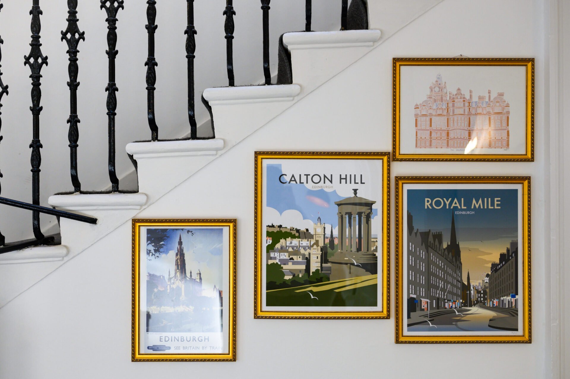 Framed travel posters on a stair wall depict Edinburgh scenes: a castle, Calton Hill, and the Royal Mile. The posters are bordered in gold frames. Black wrought iron railing is visible on the left.