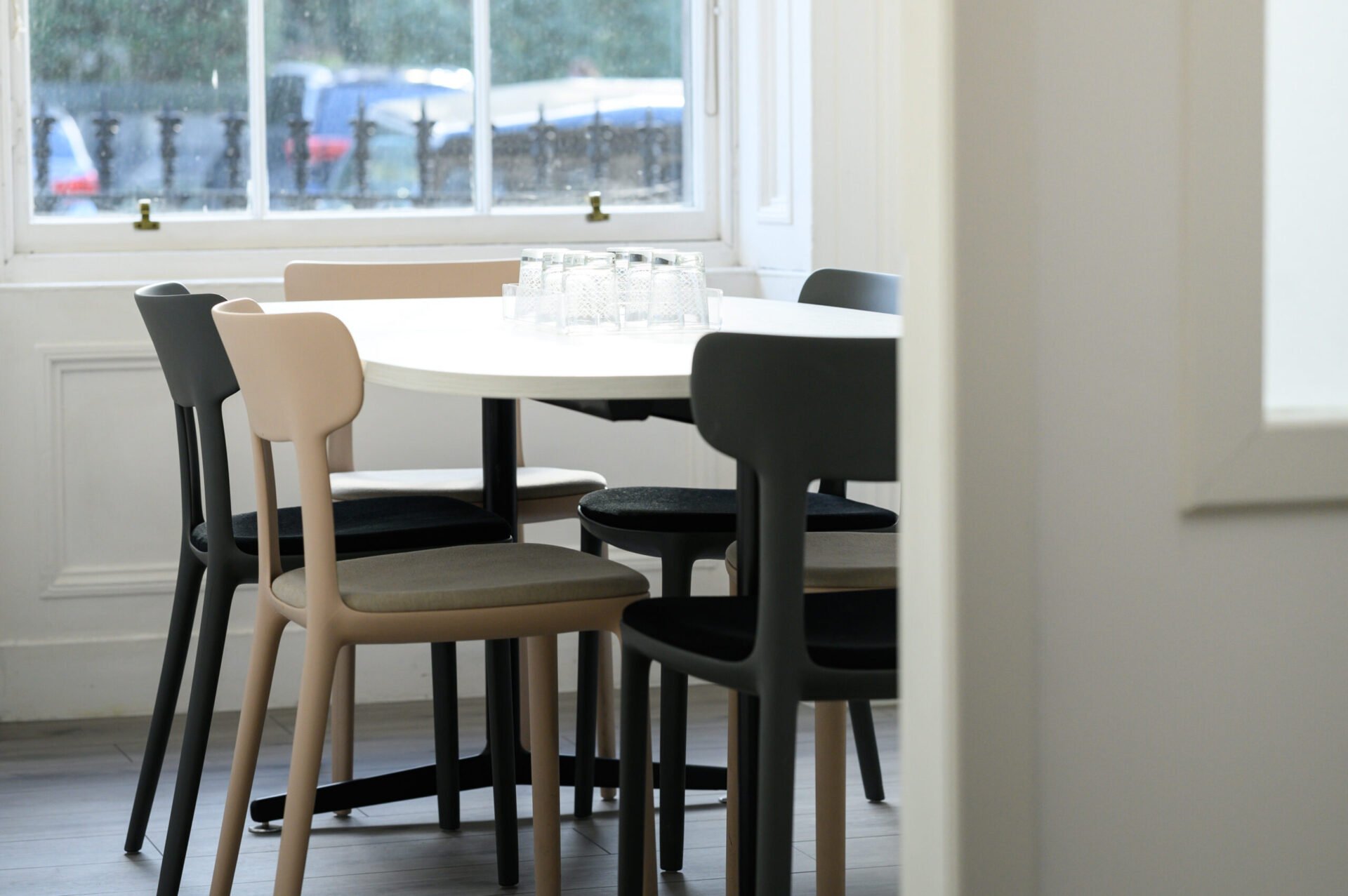 A cozy dining area with a round white table and mixed-color chairs near a window. A stack of glass cups sits on the table. The room features white walls and wooden flooring, with a view of parked cars and railings outside.
