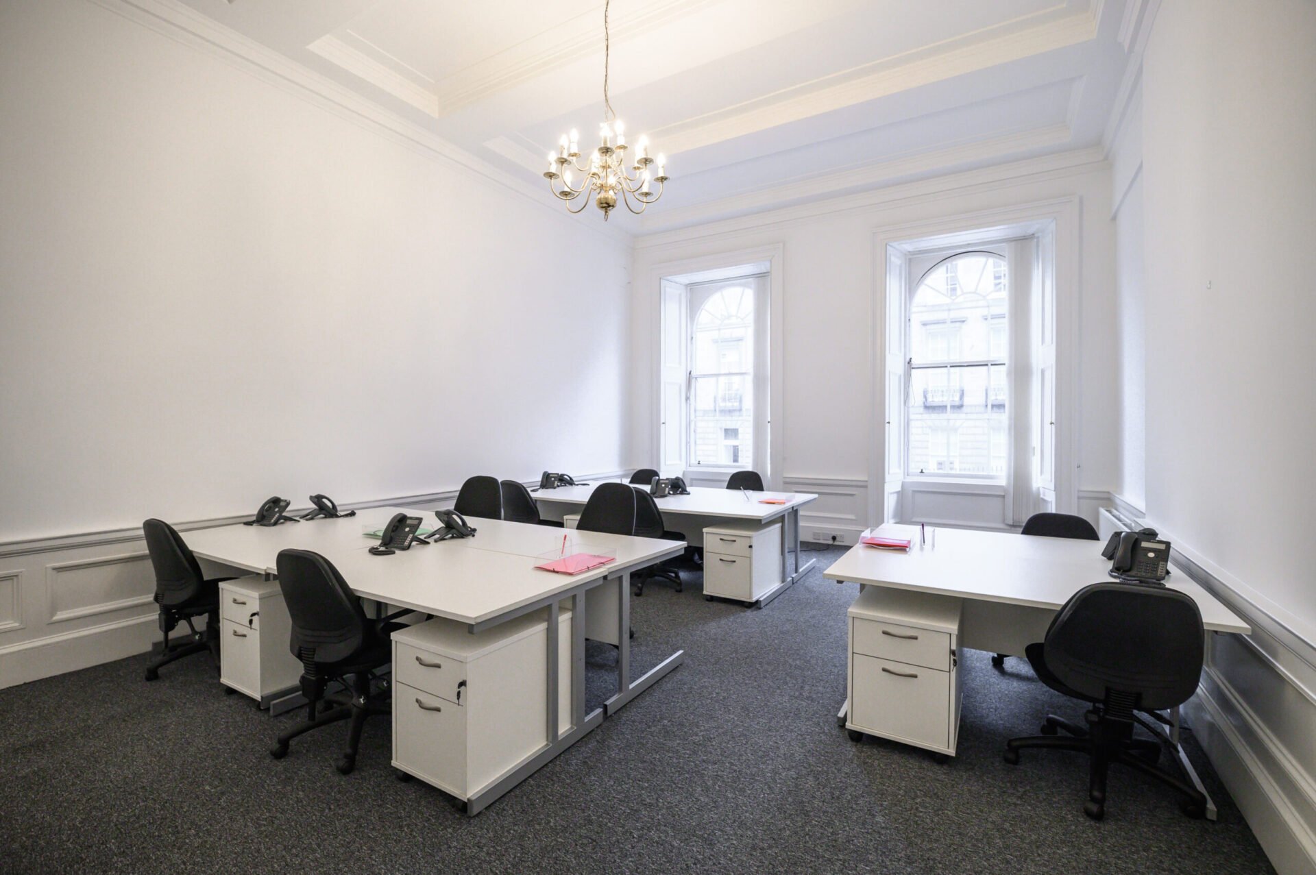 A bright, spacious office with five white desks, each equipped with a phone and office chair. The room features high ceilings, two large arched windows, and a chandelier. The floor is carpeted in gray.
