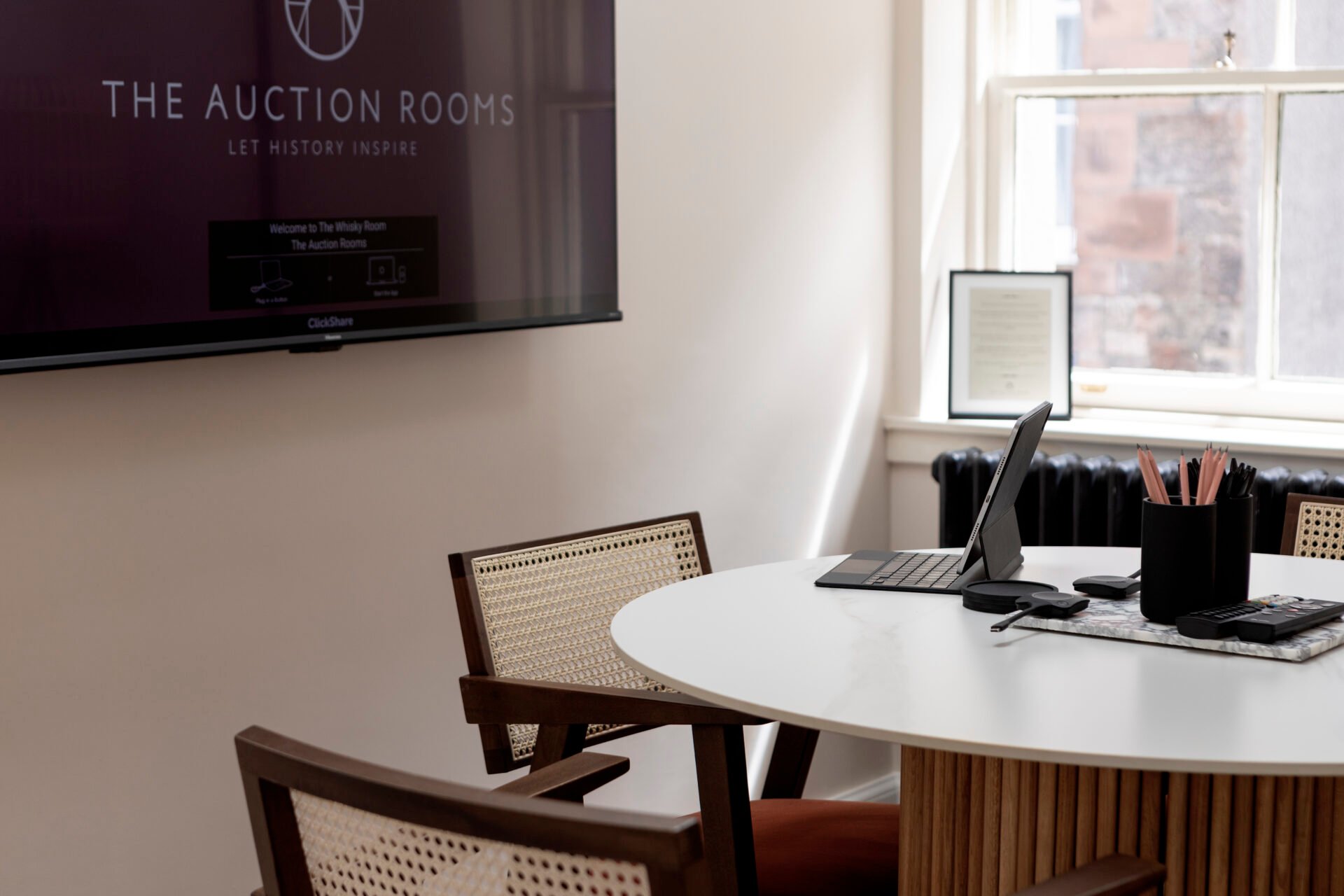 A modern meeting room at 22 Queen Street features a round table, wicker chairs, and a tablet. A sign reading The Auction Rooms, Let History Inspire adorns the wall. Bathed in natural light from a large window, the space boasts minimalistic and contemporary decor.