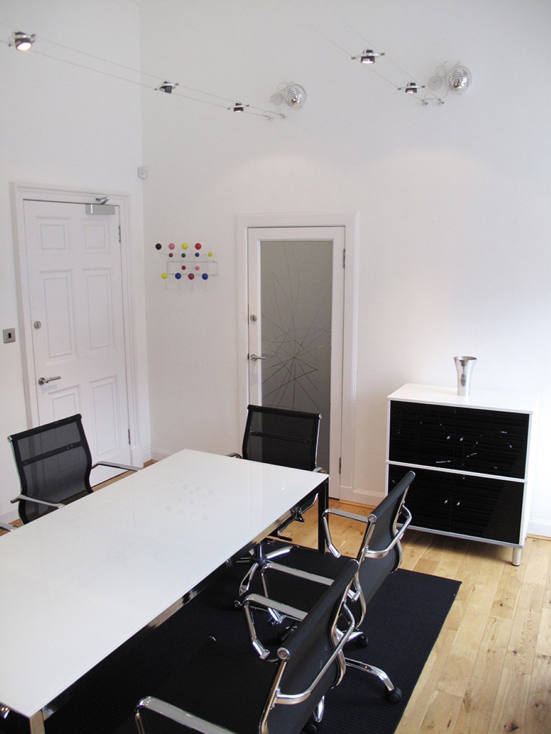 A modern office for 44 Unlimited features a white table with four black chairs and a sleek black cabinet. The room boasts wooden flooring, white walls, and a frosted glass door with a starburst pattern. Track lighting illuminates the space, while colorful dots bring life to one wall.