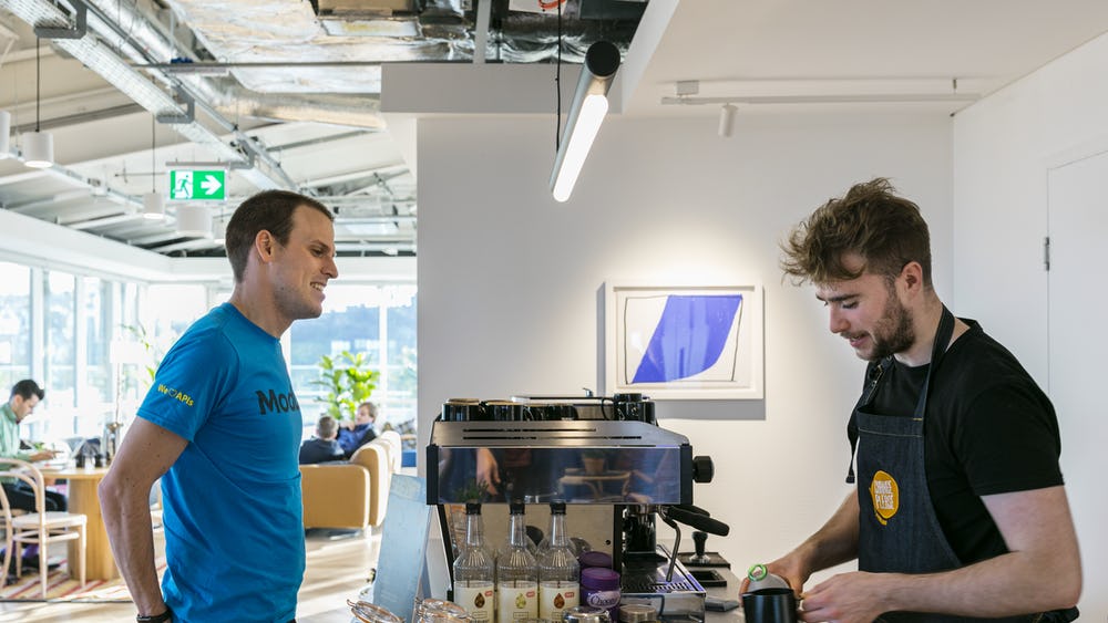 At 80 George Street, a barista in an apron crafts a drink at the coffee machine, while a man in a blue T-shirt waits at the counter in the modern cafe. The room is bright with natural light and charming seating areas are visible in the background.