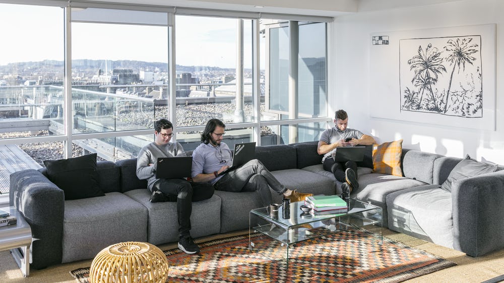 Three people are seated on a large gray sectional sofa in a bright room at 80 George Street, each working on laptops. In front of them, a glass coffee table rests on a patterned rug, while a framed artwork adorns the wall. The floor-to-ceiling windows flood the space with natural light.