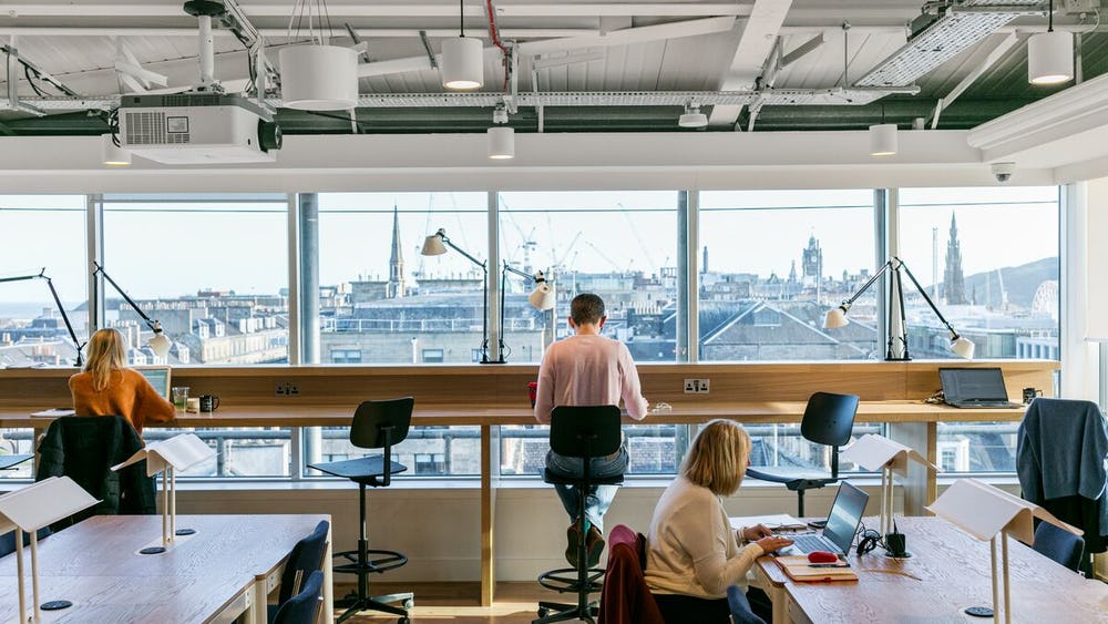 People working on laptops in a modern office at 80 George Street, featuring large windows with a city view. The room boasts high ceilings, exposed beams, and various desk lamps. Natural light fills the space, creating a bright and open atmosphere.