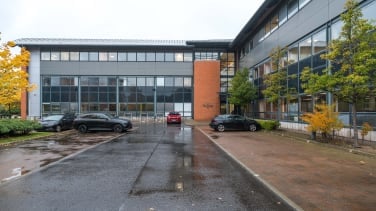 Modern office building with large windows, gray and brick exterior, at South Gyle. In front, a wet parking lot with three cars under a cloudy sky. Sparse trees with yellow leaves line the side, suggesting autumn.