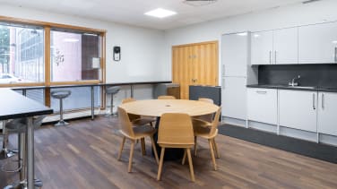 Modern kitchen with wooden flooring, round table, and five wooden chairs. White cabinets with a gray countertop and sink. Large window offering a South Gyle view, high stools along a counter. Bright lighting and minimalist design.