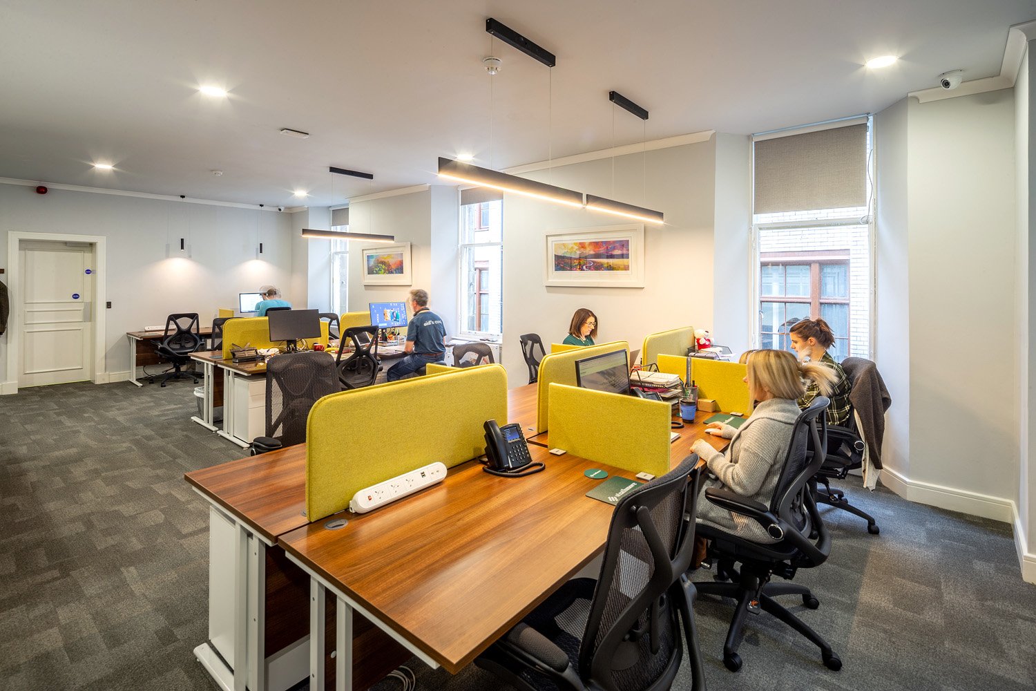Scott House features a modern office space bustling with activity. Wooden desks, yellow dividers, and black chairs populate the room, while large windows usher in natural light. Framed artwork adds flair to the walls, and overhead lighting ensures every corner is well-illuminated.