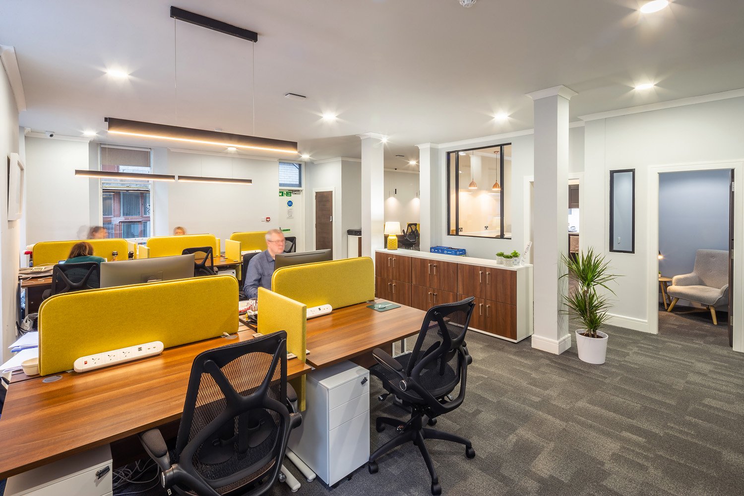 The modern open-plan office at Scott House features wooden desks and yellow dividers. Ergonomic black chairs are positioned at each workstation, with a potted plant adding a touch of nature. A corridor leads to another area, while natural light floods the space, enhancing productivity.